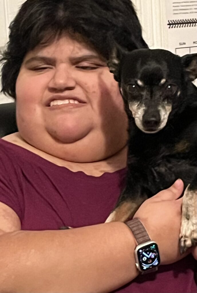 A light-skinned woman with a round face, short black hair, eyes half-closed, is smiling while holding a little black dog with a mostly white muzzle up near her shoulder. The dog is looking straight at us with a very intent look on its face.